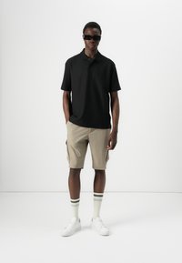 Svart polo-skjorte, beige cargo-shorts, hvite joggesko og grønstripe sokker. Modell står mot en ensfarget bakgrunn, med hendene i lommene.
