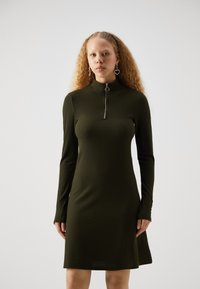 Olivově zelené šaty s ribovým vzorem, dlouhými rukávy a límcem na poloviční zip. Tkanina vypadá měkce a pružně, s přiléhavým siluetou.