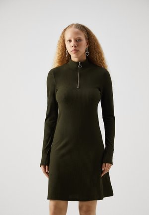 Robe côtelée vert olive avec des manches longues et un col zippé à mi-hauteur. Le tissu semble doux et extensible, avec une silhouette ajustée.