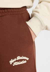 Bruine sweatpants van zacht materiaal met een elastische tailleband, voorzien van een zijzak en witte "New Balance Athletics" logo detail.