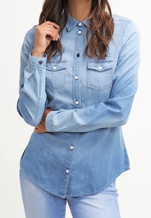 Kvinde iført en lyseblå denimskjorte med knapper og brystlommer med klap, stående med armene krydset og den ene hånd tæt ved ansigtet.