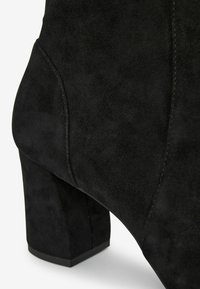 Bottine en daim noir avec un talon épais. Présente une texture lisse, un design courbé et des détails de couture subtils le long des côtés.