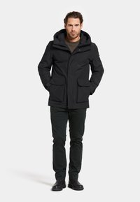 Svart vattentät jacka med huva, framknäppning och två stora framfickor. Kombinerad med mörka byxor och svarta stövlar.