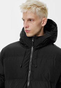 Črna puhasta jakna z visokim ovratnikom in kapuco, z zadrgo spredaj in vrvicami. Mat tekstura, prešita zasnova in elegantna silhueta.