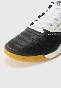 Svart innendørs fotsko for soccer med en glatt tekstur, polstret tunge, hvite lisser og såle av gummigummi. Nøkkeldetaljer inkluderer ventilasjonsakenter.