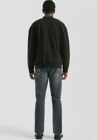 Man in een zwart langemouwen shirt, vervaagde grijze jeans en zwarte schoenen, staand met zijn rug naar de camera tegen een effen lichtgrijze achtergrond.