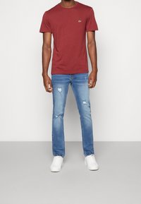 Maroon crew neck t-shirt with small green logo, paired with light blue distressed jeans and white sneakers. Casual fit, cotton blend fabric.