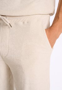 Ljusbeige joggers i mjuk terrytjänst har en elastisk midja med dragsko och sidofickor, vilket visar en texturerad, plysch yta.