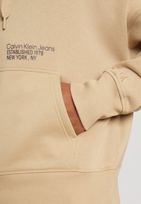 Felpa beige in cotone con tasca a marsupio. Presenta il logo ricamato "Calvin Klein Jeans" e "CK" sulla manica. Tessuto morbido e polsini a coste.
