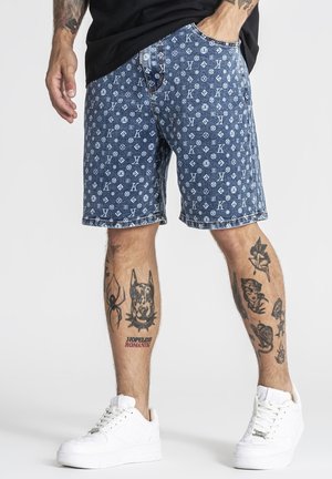 Mand iført blå mønstrede shorts med initialer, hvide sneakers og tatoveringer, herunder en edderkop, et hundehoved og sætninger på benene.