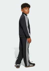 Young boy standing sideways, wearing black sportswear with white stripes and black Adidas sneakers with white soles.