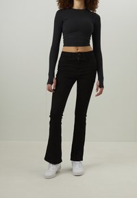 Personne portant un crop top noir à manches longues, un pantalon flare noir taille haute et des baskets blanches, debout devant un fond clair uni.