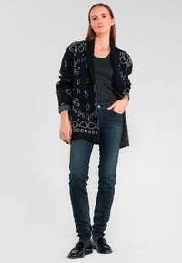 Cardigan noir avec des motifs circulaires blancs, à ouverture frontale et à manches longues. Porté sur un haut sombre et associé à un jean et des chaussures sombres.