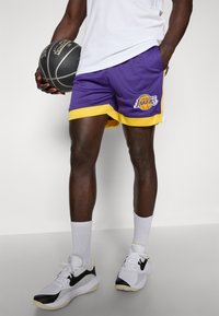 Șorturi de baschet din plasă, violet și galben, cu accente laterale, având logo-ul Los Angeles Lakers. Purtați cu un tricou alb și o minge de baschet.