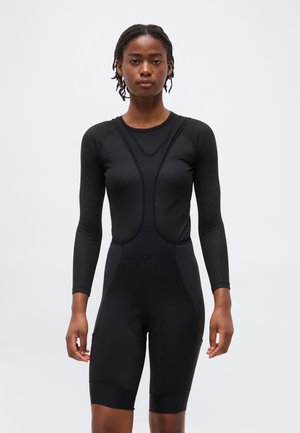 Femme portant un cuissard de cyclisme noir à manches longues ajusté, debout, face à un fond uni.