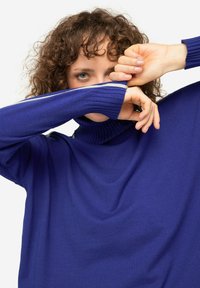 Maglione blu royal con collo alto, dalla texture a coste e maniche lunghe. Le braccia del modello coprono parzialmente il viso, enfatizzando il design del capo.