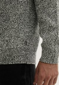 Grijze en witte gebreide trui met een textuurpatroon, geribbelde manchetten en een klein logo-detail nabij de zoom.