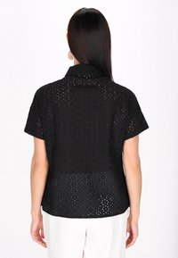 Maglietta nera a maniche corte con motivo ad occhio tagliato. Presenta un colletto e una vestibilità rilassata. Il materiale sembra leggero e tessurizzato.
