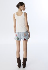 Canottiera bianca a costine abbinata a pantaloni corti di jeans azzurri chiari con colorate ricami di frutta sulle tasche posteriori e stivali marroni morbidi.