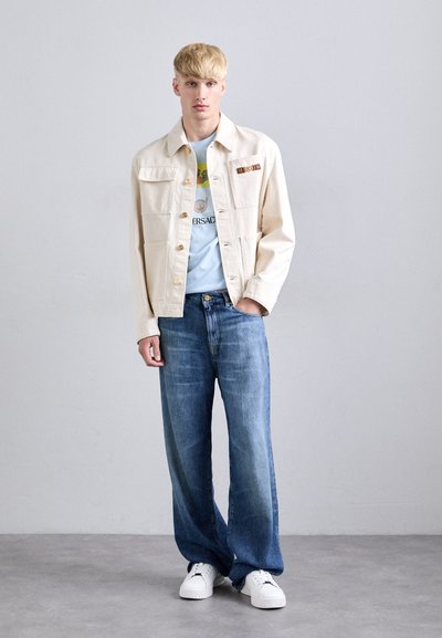 Young man with blonde hair wearing a cream jacket, light blue graphic t-shirt, loose blue jeans, and white sneakers standing against a plain wall.