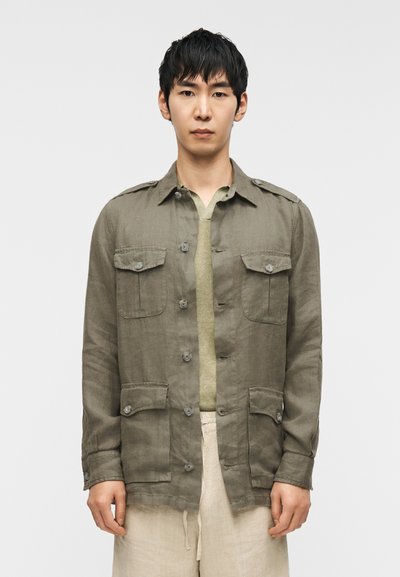 Veste en lin kaki avec quatre poches frontales, fermeture par boutons et épaulettes. Portée sur un haut vert et un pantalon beige.