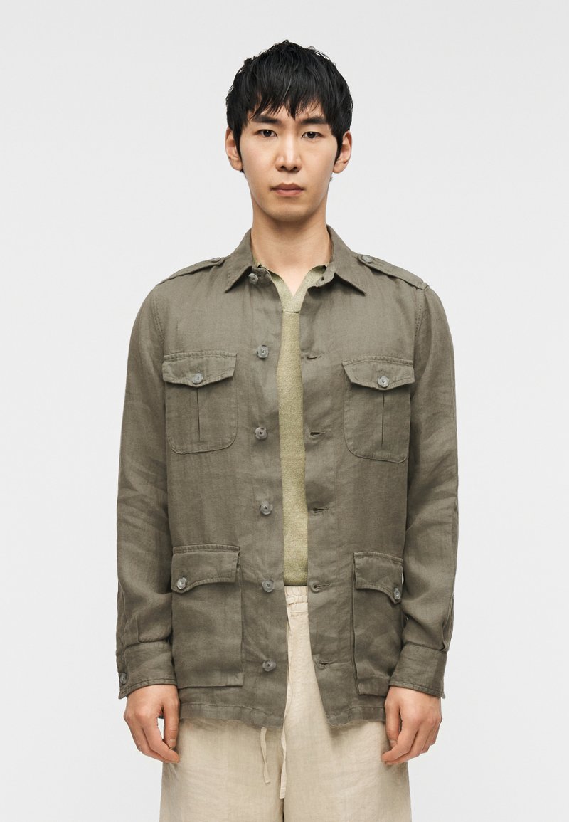 Χακί λινό σακάκι με τέσσερις τσέπες μπροστά, κλείσιμο με κουμπιά και επιθεωρήτες στους ώμους. Φορεμένο πάνω από πράσινο τοπ και μπεζ παντελόνι.