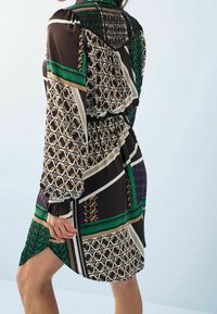 Mujer que lleva un vestido de longitud a la rodilla con un patrón de diseños geométricos en verde, negro, beige y púrpura, de mangas largas y con la cintura ajustada.