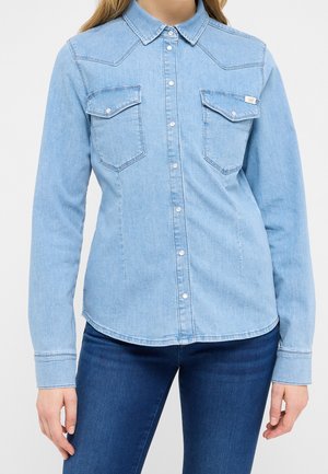 Kobieta ubrana w jasnoniebieską jeansową koszulę na guziki z dwoma kieszeniami na piersi oraz ciemnoniebieskie jeansy, stojąca na jednolitym tle.