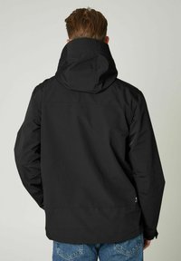 Czarna wodoodporna kurtka z kapturem, o gładkiej fakturze, z zapięciem na zamek błyskawiczny z przodu i elastycznymi mankietami. Noszona na niebieskie dżinsy.