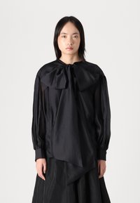 Zwarte blouse met een grote strik bij de hals, doorschijnende lange mouwen en een textuurstof. Heeft een asymmetrische zoom en geplooide accenten.