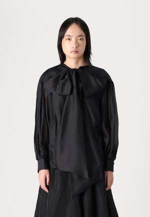 Svart bluse med en stor sløyfe ved halsen, gjennomsiktige lange ermer og strukturert stoff. Har en asymmetrisk underkant og plisserte detaljer.
