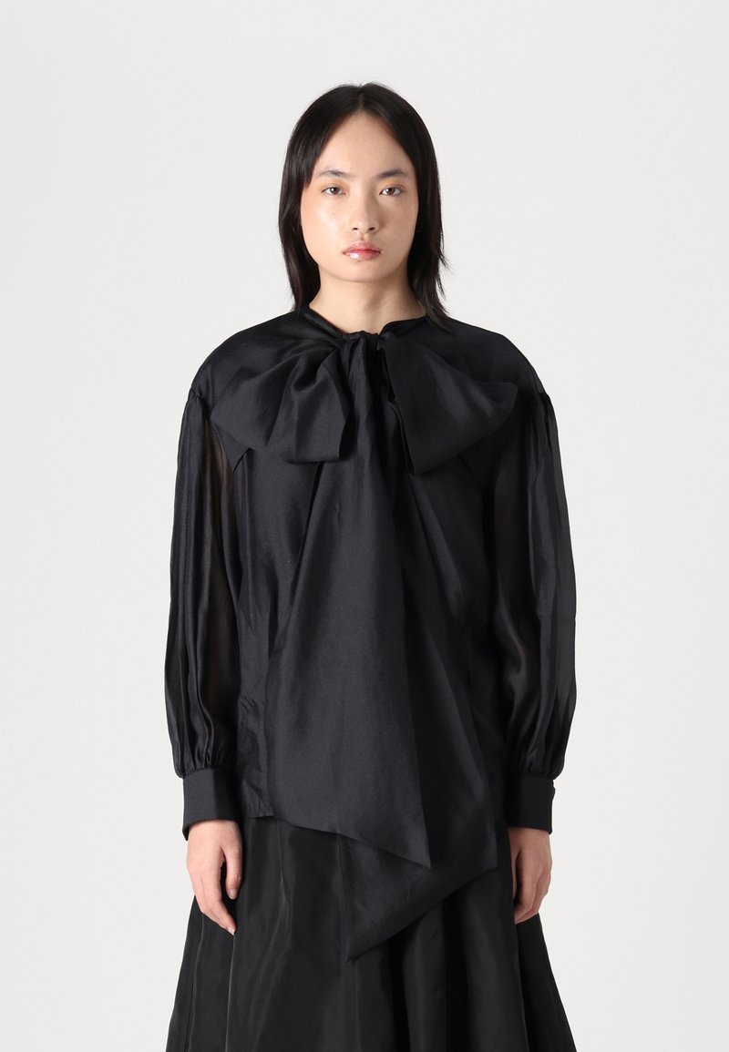 Zwarte blouse met een grote strik bij de hals, doorschijnende lange mouwen en een textuurstof. Heeft een asymmetrische zoom en geplooide accenten.