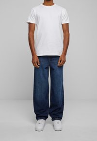 Person som bär en enfärgad vit t-shirt, lösa mörkblå jeans och vita sneakers, stående mot en ljusgrå bakgrund.