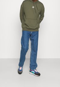 Sudadera con capucha verde con bolsillo canguro y logo blanco, vaqueros azules de pierna recta y zapatillas multicolor con detalles en contraste.