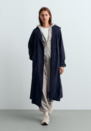 Jeune femme marchant vers l'avant portant un long manteau bleu marine sur un pantalon de survêtement gris, un sweat à capuche gris zippé, une chemise blanche, des chaussettes blanches et des baskets beiges.
