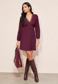 Burgundi színű ruha hosszú puff ujjakkal és csipkével díszített nyakkivágással, összeillő térd kérgű csizmával és díszített táskával párosítva.