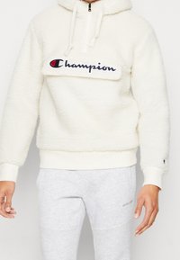 Champion krämfärgad fleecehoodie med halvdragkedja, broderad logotyp på bröstet, ribbade ärmslut och fåll, med en mjuk, texturerad finish.