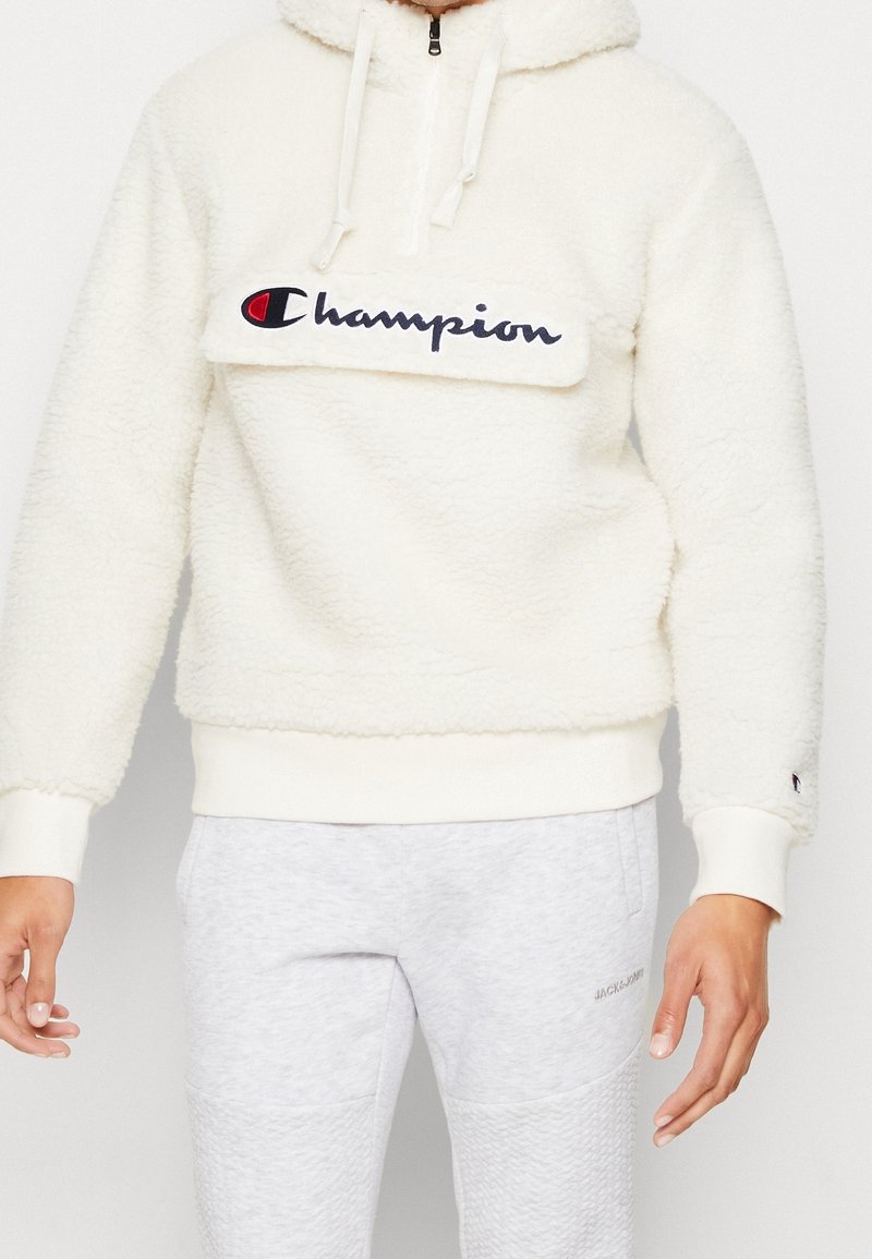 Champion krämfärgad fleecehoodie med halvdragkedja, broderad logotyp på bröstet, ribbade ärmslut och fåll, med en mjuk, texturerad finish.