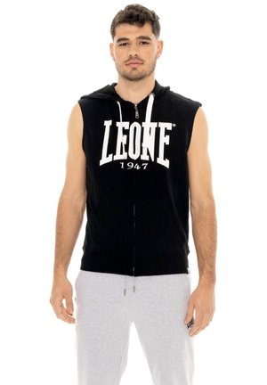 Un homme porte un sweat à capuche noir sans manches, zippé et orné d’une inscription blanche, ainsi qu’un pantalon de jogging gris clair, sur fond blanc.