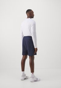 Pantalones cortos azul marino con una cintura elástica, combinados con una blusa blanca de manga larga. La modelo está de perfil, mostrando un diseño minimalista.