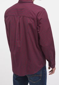 Camisa de cuadros roja y azul marino con botones, mangas largas, pliegue trasero y dobladillo redondeado, combinada con pantalones vaqueros azules.