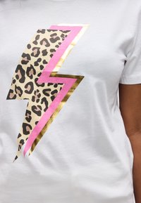 Λευκό πουκάμισο με μεγάλο γραφικό κεραυνού που περιλαμβάνει μοτίβο λεοπάρδαλης, ροζ και μεταλλικές χρυσές αποχρώσεις στο μπροστινό μέρος.