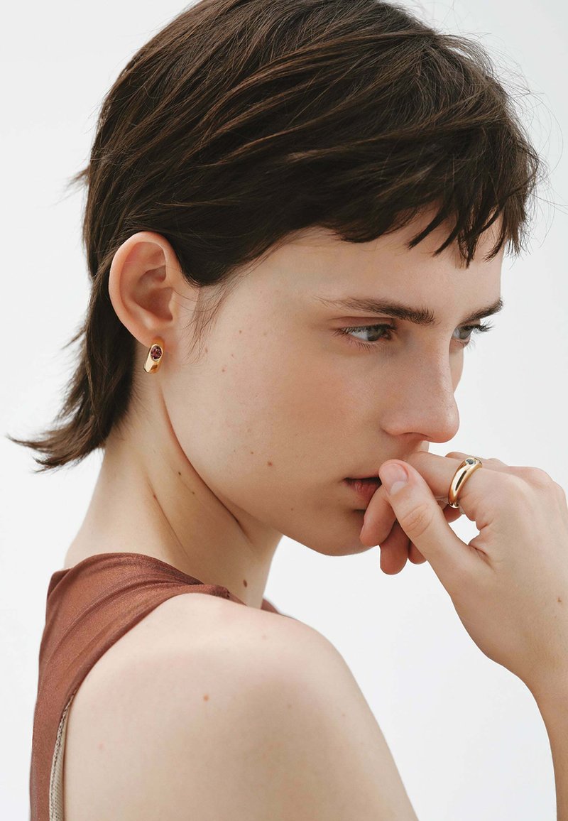 Pendiente de aro dorado con un detalle de gema roja, llevado por una modelo. Ella tiene el cabello corto y oscuro, lleva una blusa marrón, con los dedos cerca de sus labios.