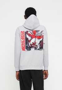 Γκρι φούτερ με κουκούλα, με μεγάλο γραφικό που απεικονίζει ένα πρόσωπο σε αποχρώσεις κόκκινου και γκρι, και το κείμενο "METAL GEAR" με έντονα κόκκινα γράμματα.