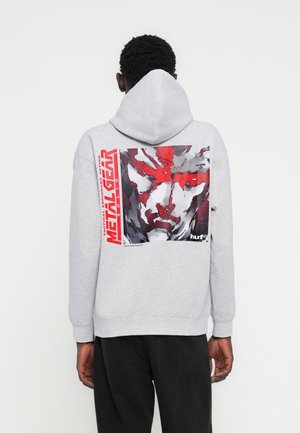 Γκρι φούτερ με κουκούλα, με μεγάλο γραφικό που απεικονίζει ένα πρόσωπο σε αποχρώσεις κόκκινου και γκρι, και το κείμενο "METAL GEAR" με έντονα κόκκινα γράμματα.