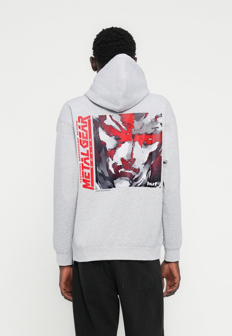 Γκρι φούτερ με κουκούλα, με μεγάλο γραφικό που απεικονίζει ένα πρόσωπο σε αποχρώσεις κόκκινου και γκρι, και το κείμενο "METAL GEAR" με έντονα κόκκινα γράμματα.