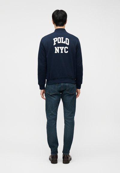 Άνδρας με σκούρο μπλε μπουφάν με λευκό κείμενο «POLO NYC» στην πλάτη, μπλε τζιν και σκούρα καφέ παπούτσια, που στέκεται με το πρόσωπο προς ένα λευκό φόντο.