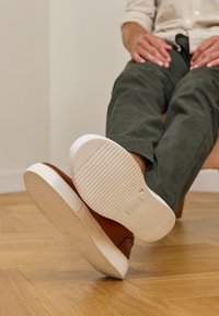 Braune Ledersneaker mit weißen Gummisohlen. Die Sohlen haben horizontale Rillen für besseren Halt. Der Hintergrund zeigt einen hellen Holzboden.