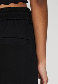 Svarta byxor med elastiskt midjeband och en mjuk textur; har en liten ficka framtill; närbild på midjan och handområdet.