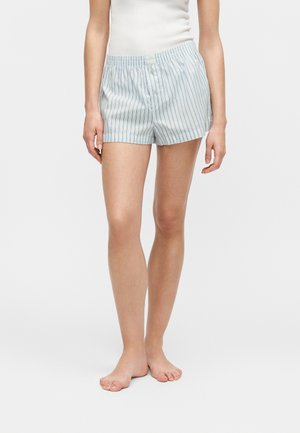Nő világoskék és fehér, függőlegesen csíkos boxer rövidnadrágban, mezítláb állva, kezei lazán oldalt.